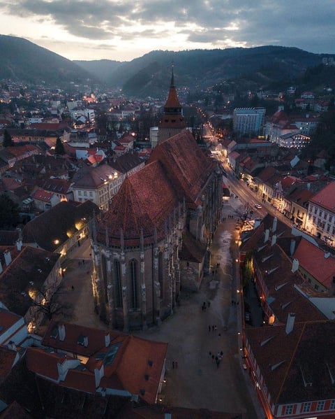 Aerial view of a Gothic church with red roof at dusk, surrounded by historic European city buildings and mountains at twilight