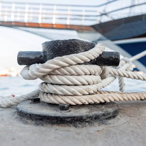 Thick white rope coiled around a black metal cleat on a dock, with boats in the background