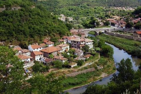 Aerial view of a riverside village nestled among green hills with traditional houses, red-tiled roofs, and a stone bridge spanning the water