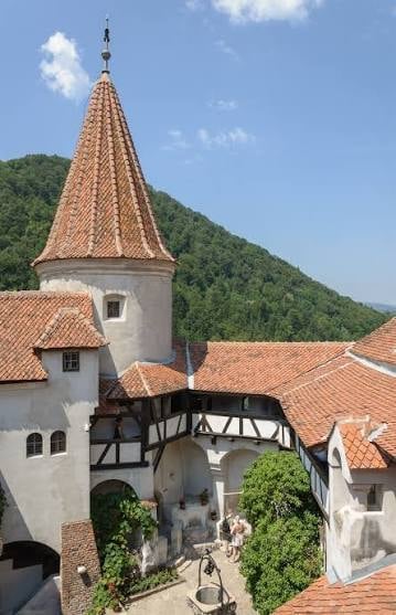 Medieval castle courtyard with conical tower, timber-frame wall, terracotta roof tiles, and mountainous forest backdrop