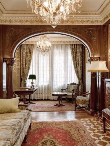 Luxurious classical bedroom with ornate chandelier, arched wooden doorway, elegant furniture, and plush carpeting in warm tones