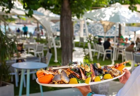 Hand holding a plate of grilled seafood and vegetables at an outdoor waterfront restaurant with white umbrellas and seating in background
