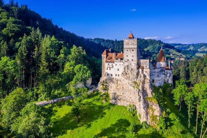Medieval castle with red-tiled roofs perched on a rocky hilltop surrounded by dense green forests and rolling mountains under a blue sky