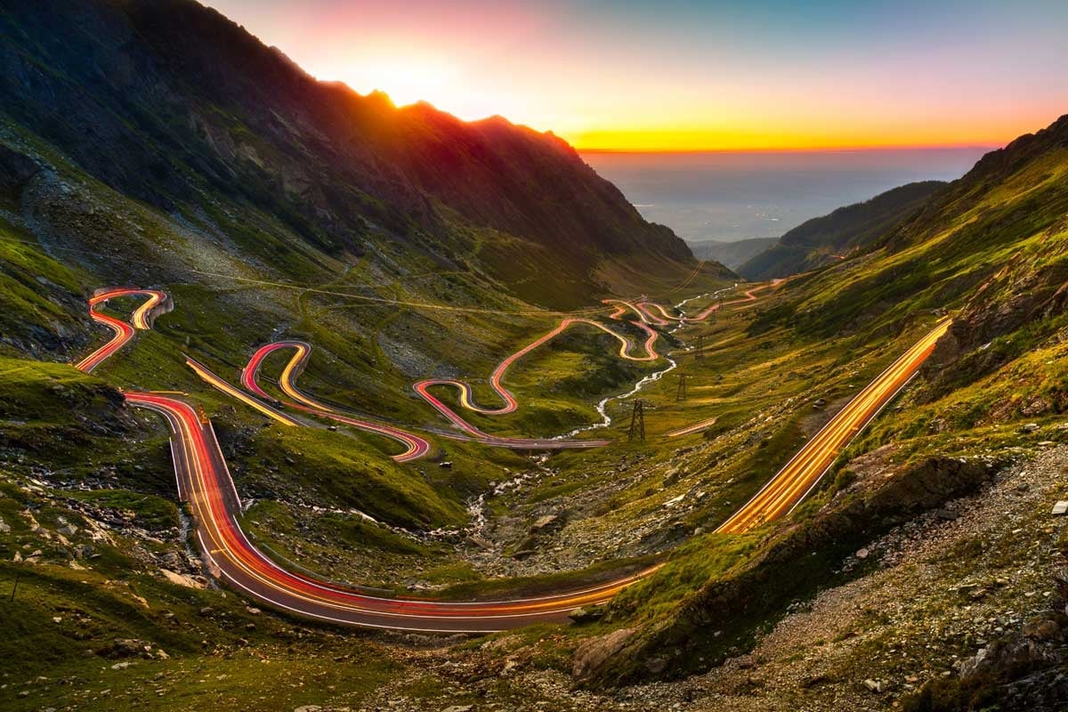 Winding mountain road with long exposure car light trails at sunset in green valley landscape