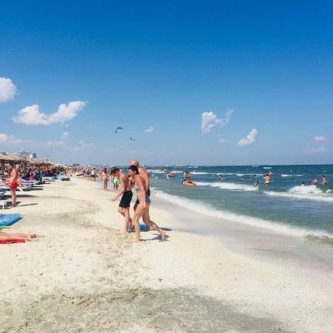Crowded beach scene with swimmers in turquoise water, sandy shore, beachgoers, and kite surfers under blue sky with white clouds