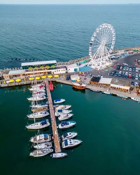Aerial view of a seaside pier with white Ferris wheel, docked fishing boats, parked cars, and beachfront attractions along turquoise water