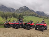 Group of people on red ATVs lined up in a mountain valley with green meadows and forested peaks in the background