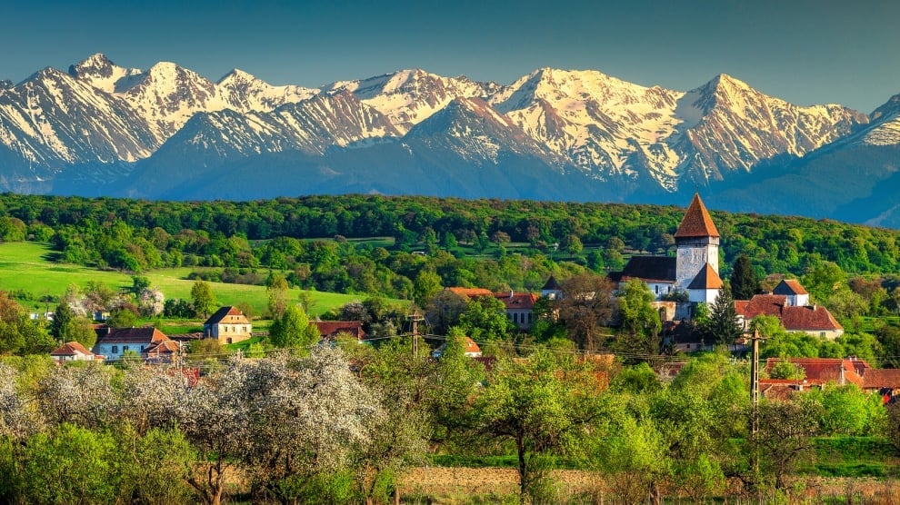 Alpine village with church nestled in green valley below snow-capped mountain range under blue sky