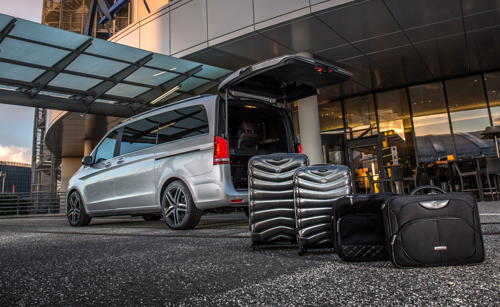 Silver minivan with open rear door displaying luggage and travel bags in front of a modern building entrance