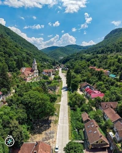 Aerial view of a small village nestled in a lush green valley with mountains, featuring a church and traditional houses with red-tiled roofs.