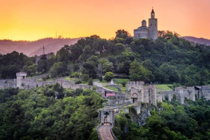 Medieval castle ruins nestled on a forested hilltop at sunset, with a church tower visible on the mountain peak beyond