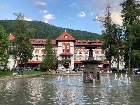 Grand historic building with red-tiled roof and tower reflected in a pond with fountain jets, surrounded by forested mountains