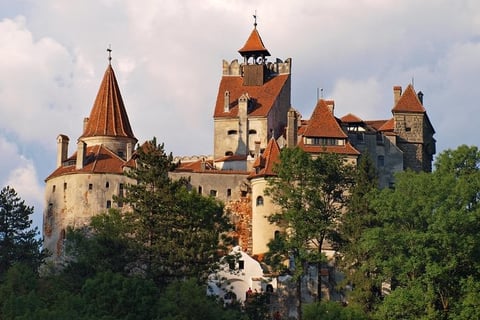 Bran Castle perched on a hilltop surrounded by green trees, featuring red-roofed towers and stone fortifications against a cloudy sky