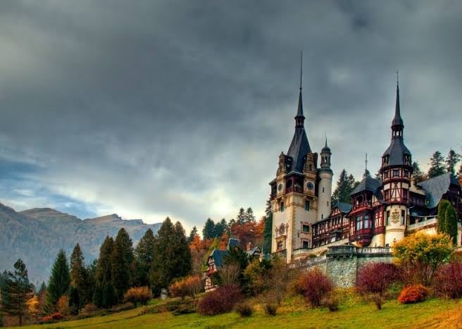 Neuschwanstein Castle with distinctive spires surrounded by autumn foliage, forests, and mountains under cloudy sky