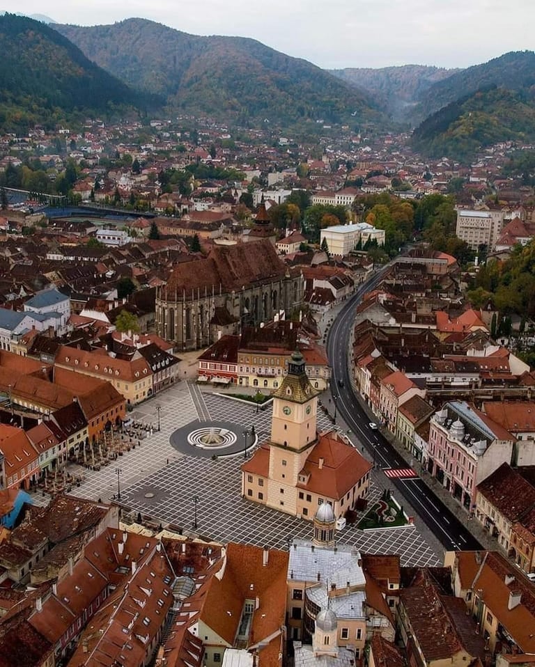 Aerial view of a medieval European city with red-tiled roofs, a town square with a clock tower, surrounded by forested mountains