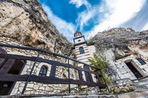 Historic lighthouse perched on rocky cliff surrounded by stone buildings and metal railings under blue sky