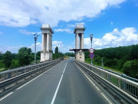 Modern bridge with twin stone towers spanning over green landscape under blue sky with white clouds