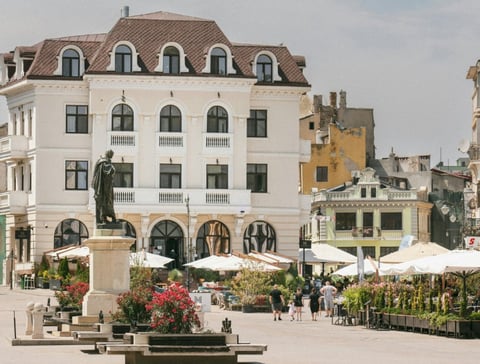 European square with white neoclassical building, statue, outdoor cafe seating with white umbrellas, and colorful historic buildings