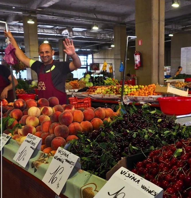 Vendor smiling at farmers market stand displaying fresh produce including peaches, leafy greens, and berries with price signs