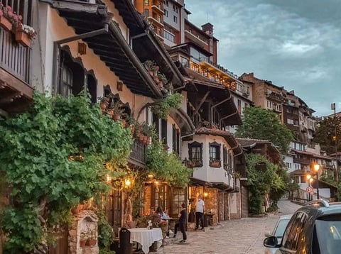 Charming European alleyway lined with ivy-covered medieval buildings, warm street lights, outdoor dining, and pedestrians at dusk