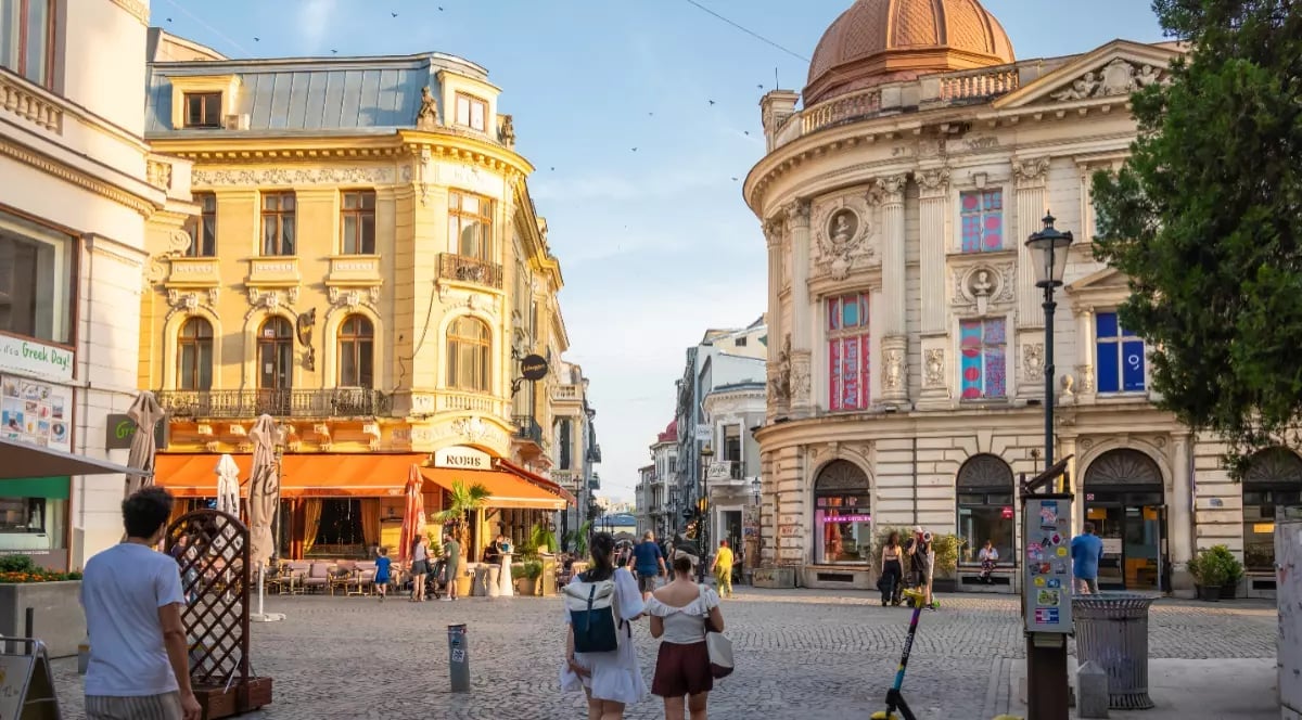 European town square with historic yellow and cream-coloured buildings, domed architecture and pedestrians walking on a cobblestone street in central Bucharest