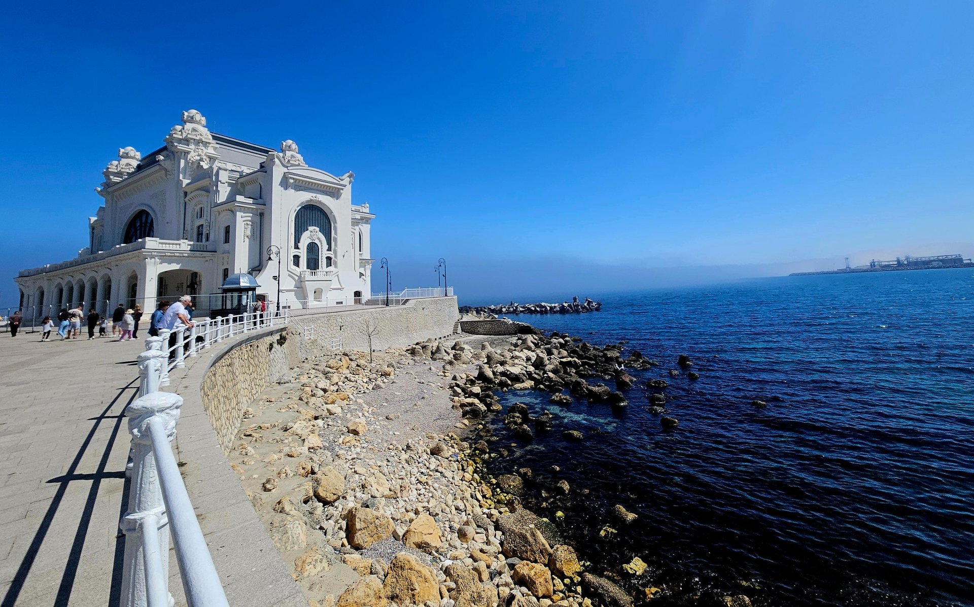 White ornate building on rocky coastal pier overlooking blue Mediterranean Sea on clear sunny day