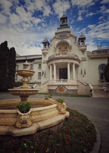 Ornate cream-colored mansion with domed tower and columned entrance, surrounded by manicured gardens and fountain courtyard under blue sky
