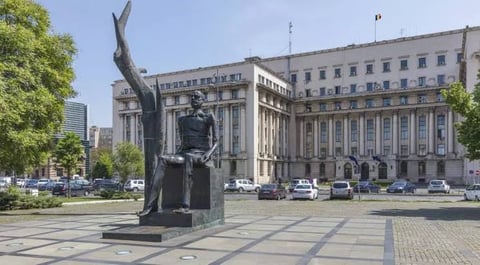 Dark statue of a seated figure in urban plaza with historic government building and trees in background