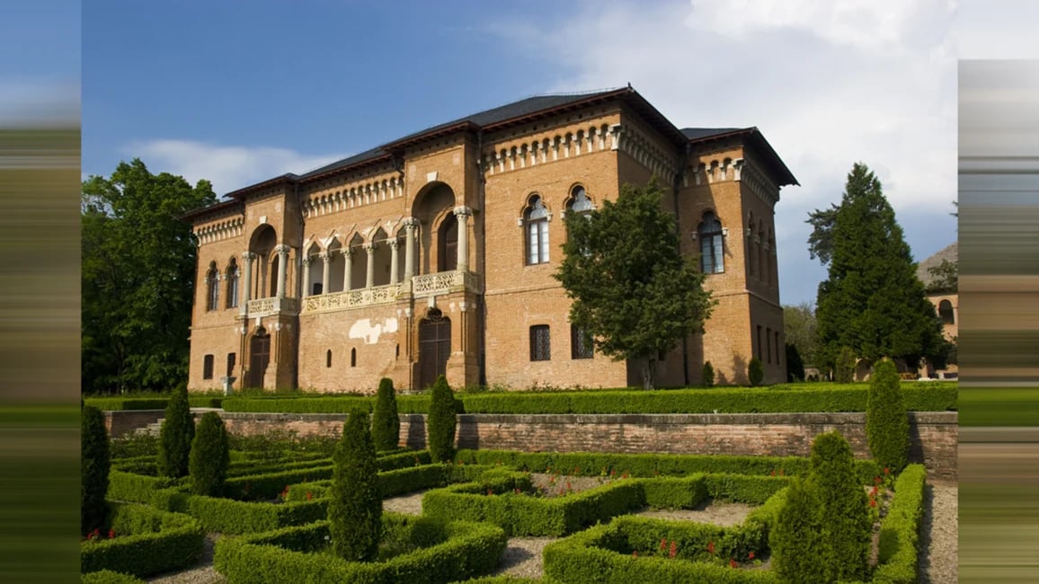 Historic European castle with ornate architecture, ivy-covered walls, formal manicured gardens with cypress hedges in foreground